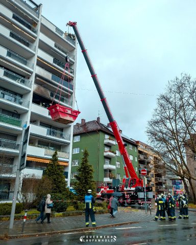 Feuerwehrkran hält eine Schuttmulde vor einen verrußten Balkon eines mehrstöckigen Hauses. Im Vordergrund stehen Feuerwehrangehörige und Passanten.